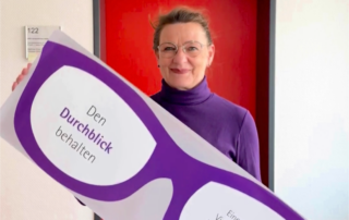 Eine Frau in lila Rollkragenpullover und Brille hält ein großes Pappschild in Form einer überdimensionalen Brille vor einem roten Türrahmen. Auf dem Schild steht: „Den Durchblick behalten“ und „Eine Initiative der Vinzenz-Konferenzen im Erzbistum Paderborn“.