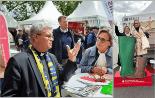 Menschen an einem Informationsstand auf einem Außenevent. Eine Person im Vordergrund spricht gestikulierend mit einer weiteren Person an einem Tisch mit Flyern. Im Hintergrund stehen Zelte mit der Aufschrift „Caritas“. Rechts: Zwei Personen stehen in übergroßen Gummistiefeln auf einem Podest mit der Beschriftung „Zusammen geht was. Caritas verbindet Generationen“.