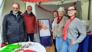 Fünf Personen stehen in einem Zelt an einem Infostand und blicken in die Kamera. Auf einem Tisch liegen Flyer und Materialien. In der Mitte steht ein gerahmtes Porträt. Rote Girlanden hängen als Dekoration im Hintergrund.