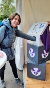 Person steht an einem Infostand im Freien und greift in eine Box mit der Aufschrift „AIC Vinzenz-Konferenz“. Die Box steht auf gestapelten Würfeln mit Logo. Auf einem Tisch liegen Karten, im Hintergrund sind Bäume und ein Zelt zu sehen. 